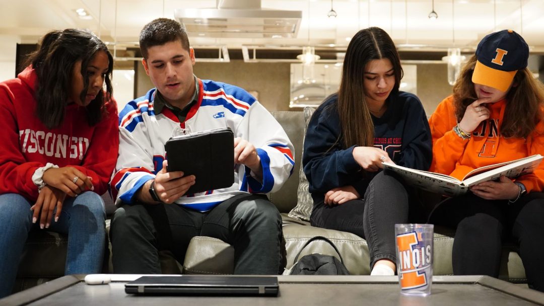 Four college students sit on a couch looking at a tablet and reading a book