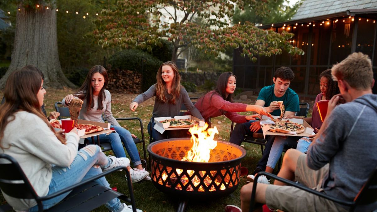 Teenagers gathering around a fire pit and eating pizza