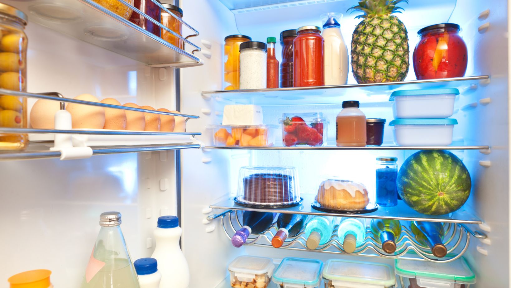Interior of a refrigerator. In the top door bin are several jars. The bin below has eggs and a jar of green olives. The bottom door bin has a glass bottle and plastic bottles of milk. The body of the fridge has four shelves containing condiments, produce, two cakes and several bottles of wine.