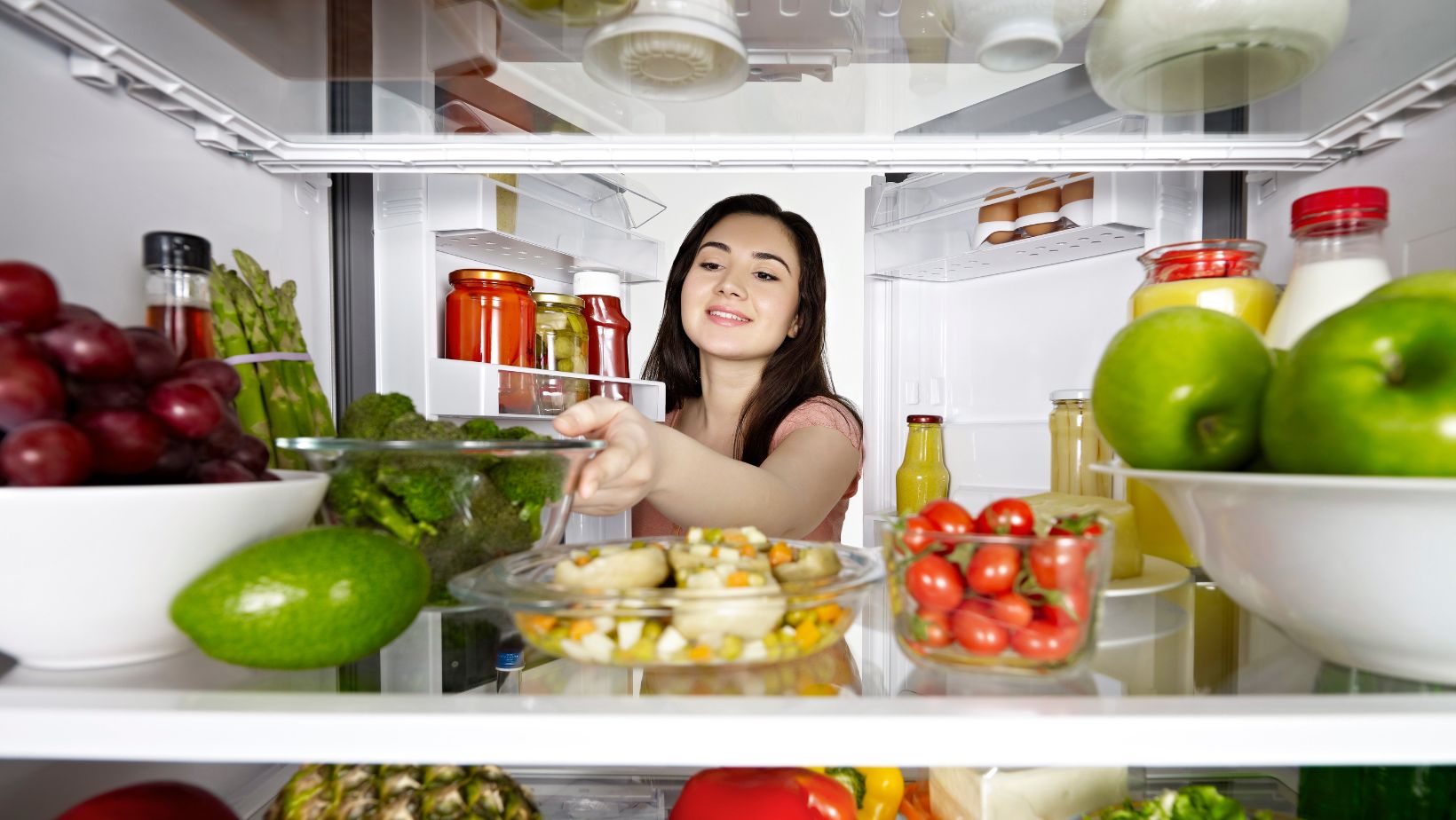 woman reaching into the interior of a refrigerator. different fridge organization methods are on display including clear organizing bins that hold produce