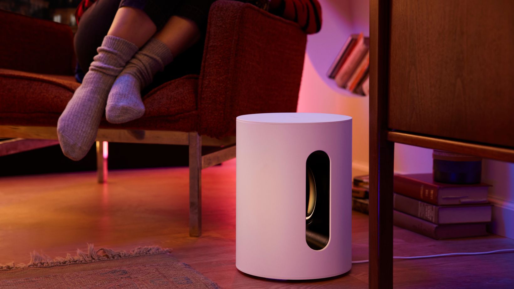 white Sonos sub mini on the floor next to an armchair. We see the grey-socked feet of the person sitting in the chair