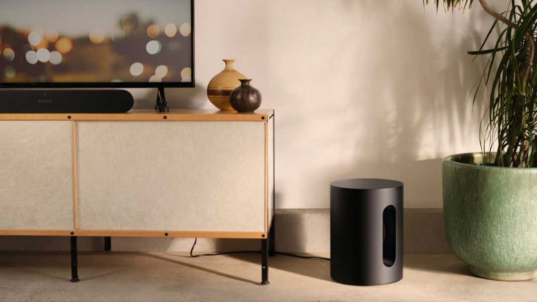 a black Sonos sub mini sits on the floor with a beige credenza to its left and a green potted plant to its right