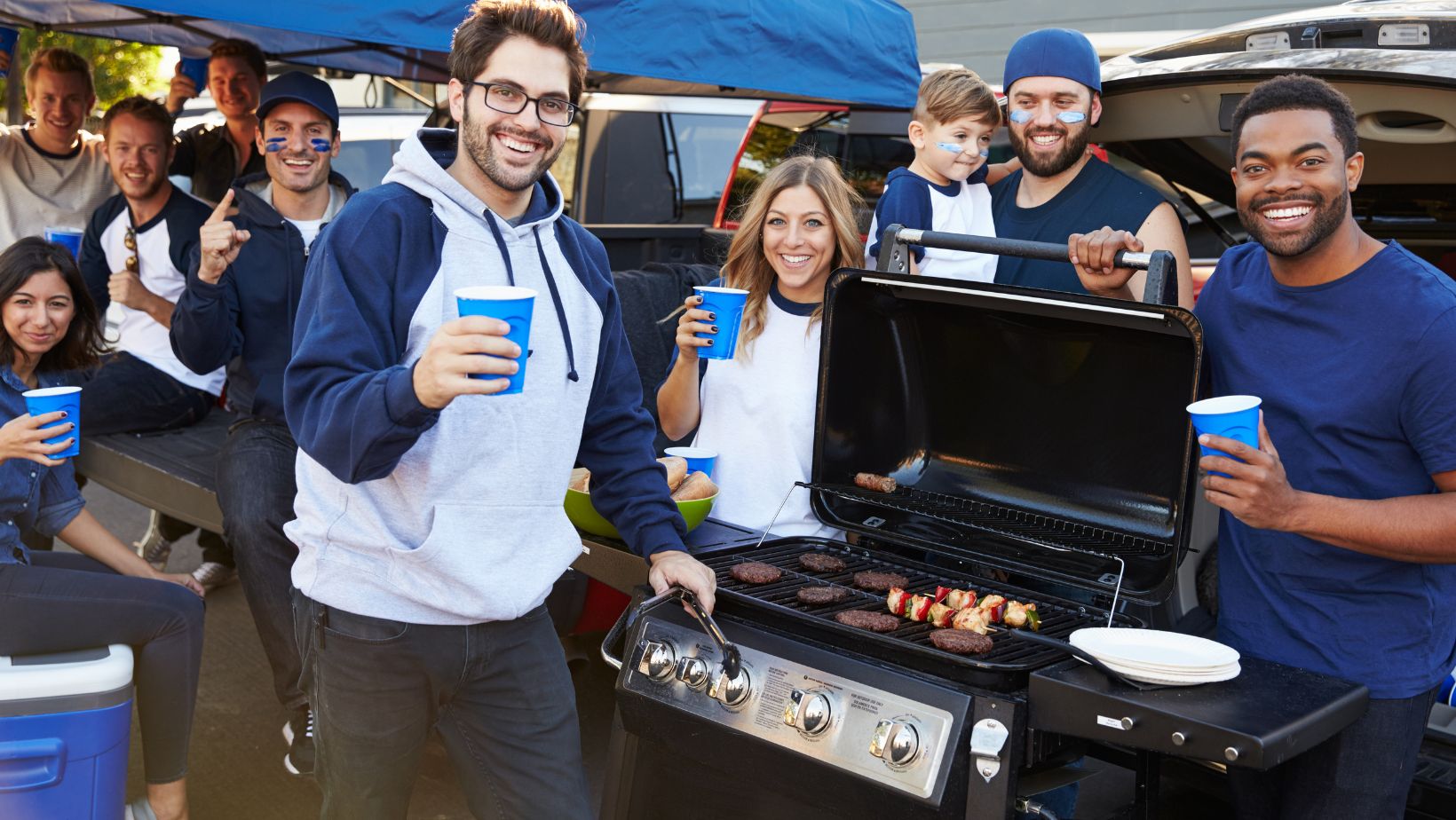 group of people wearing navy and white shirts. in the foreground, a man with brown hair standing in front of a grill holding tongs in his left hand and a blue plastic cup in his right hand