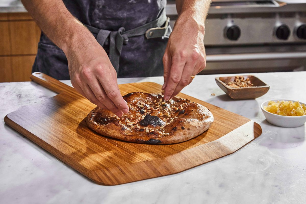 person wearing an apron sprinkling pecans and candied ginger on a pizza set on a wooden pizza peel