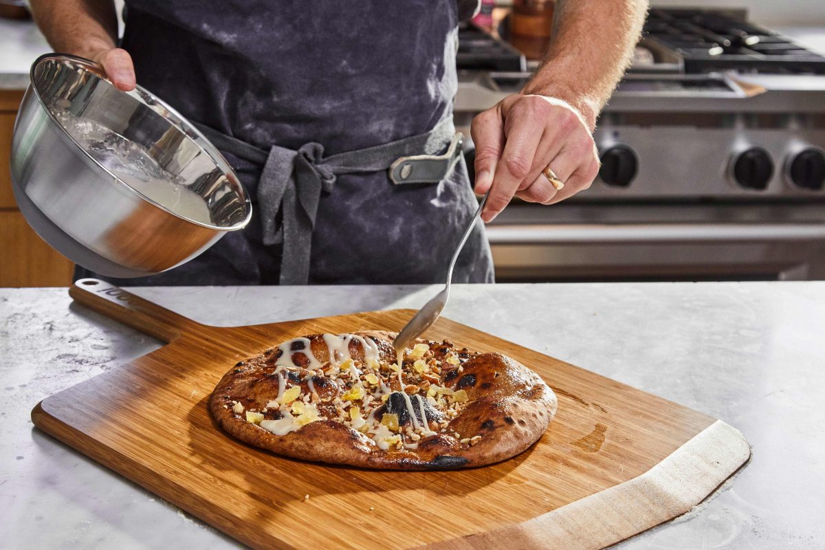 person wearing a navy apron holds a silver bowl in their left hand. With their right hand, they drizzle icing on top of a dessert pizza