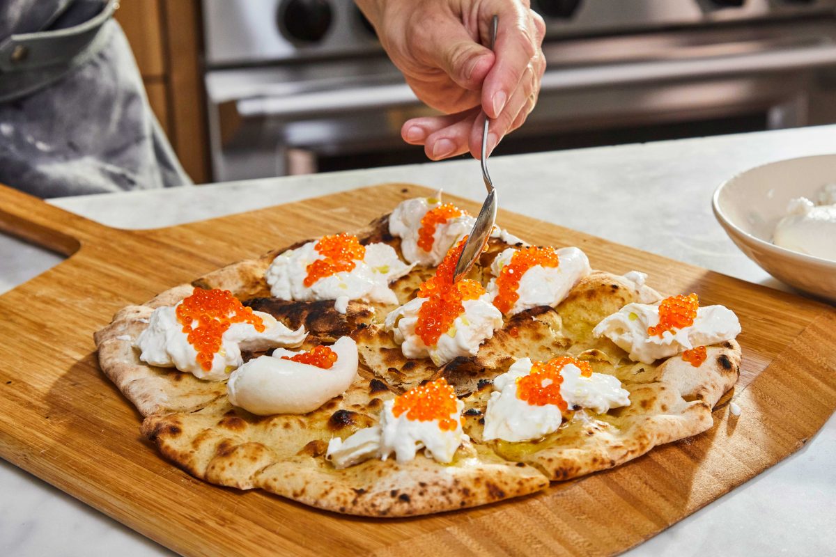 pizza crostini on a wooden pizza peel with someone spooning orange trout roe on top