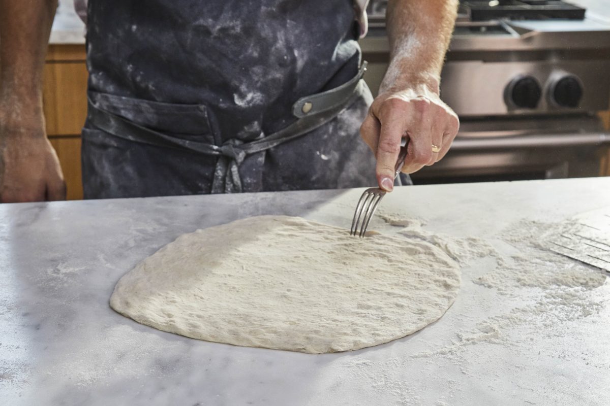 person wearing an apron docking pizza dough with a silver fork