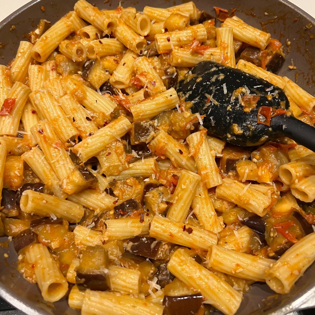 spatula stirring rigatoni noodles, eggplant and tomatoes in a pan