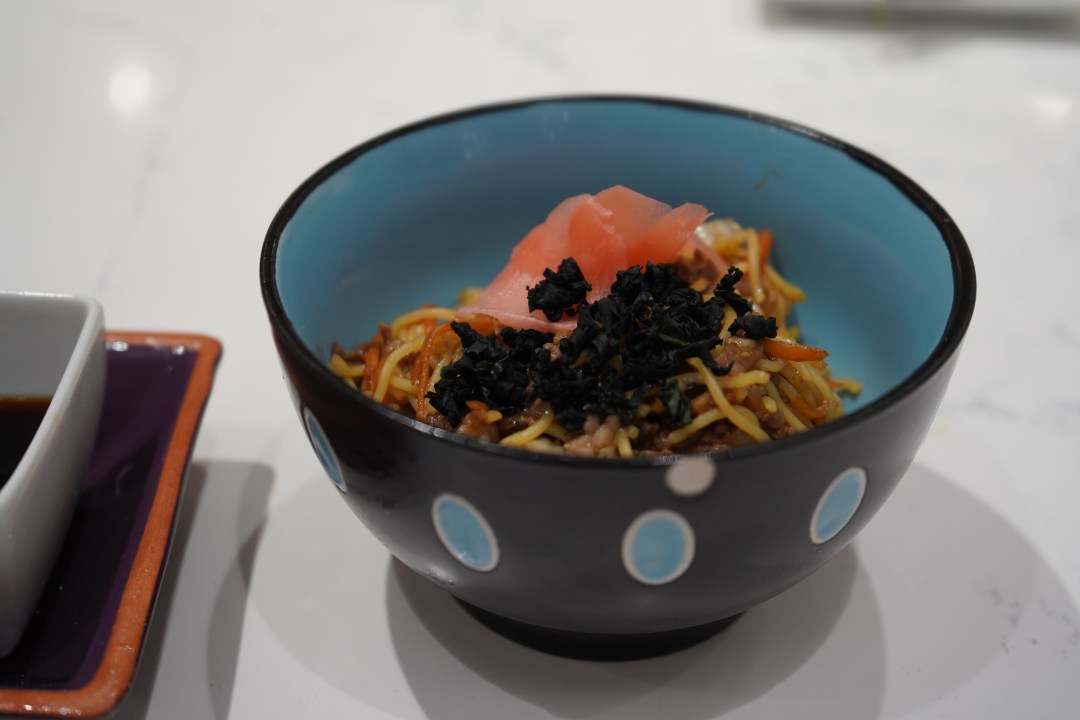 Yakisoba in a black and blue bowl topped with ginger and seaweed flakes