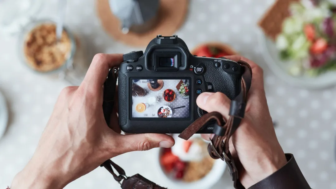 A person holding a professional camera over a table of food, taking a high quality photo that we can see in the viewfinder screen.