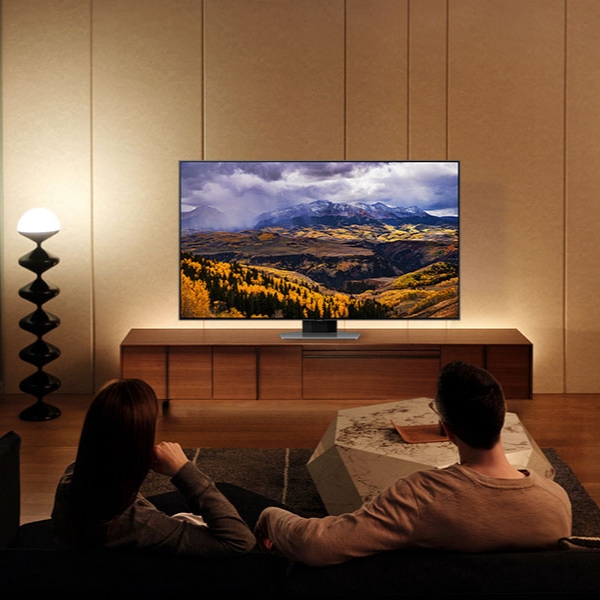 A couple sitting on a living room couch watching a Samsung Q80C TV, displaying a photo of a mountain range.
