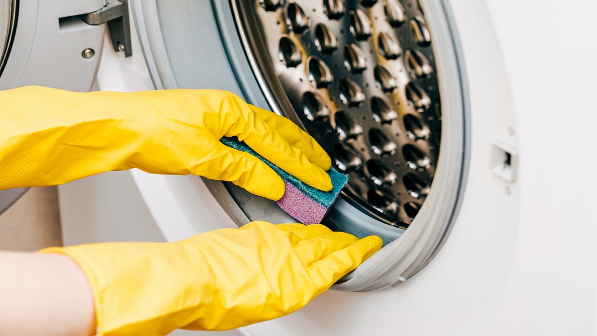 A person wearing yellow gloves cleaning a washing machine with a sponge.
