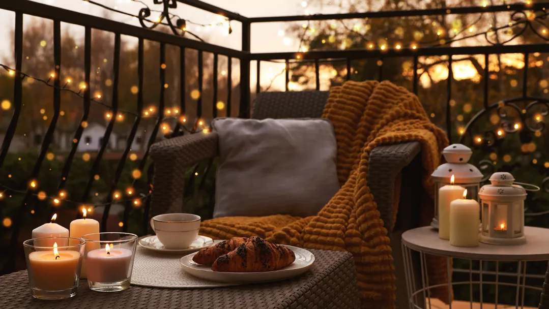 A fenced in patio with string lights, a chair, pillow and blanket. Two small tables have lit candles, lanterns, a tea cup and croissants.
