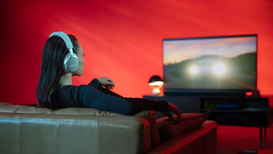A woman sits on a leather couch, watching a TV wearing the Bose QuietComfort Ultra Headphones