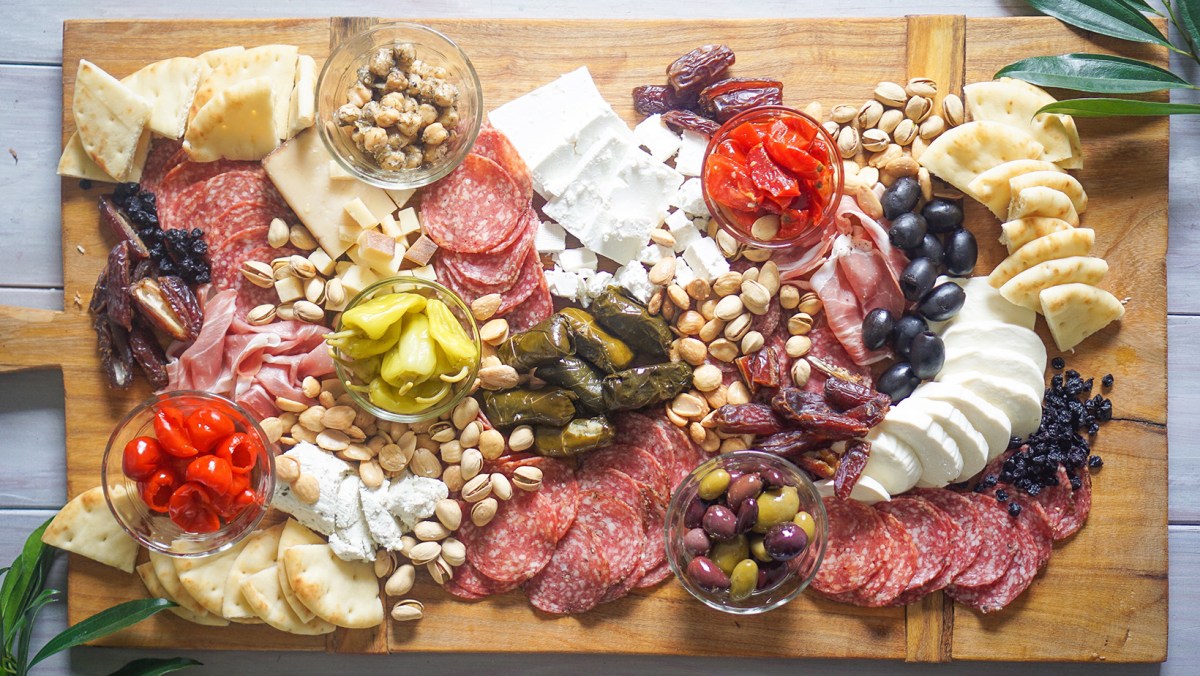 A wood cutting board covered in meats. cheeses, olives, veggies and more.