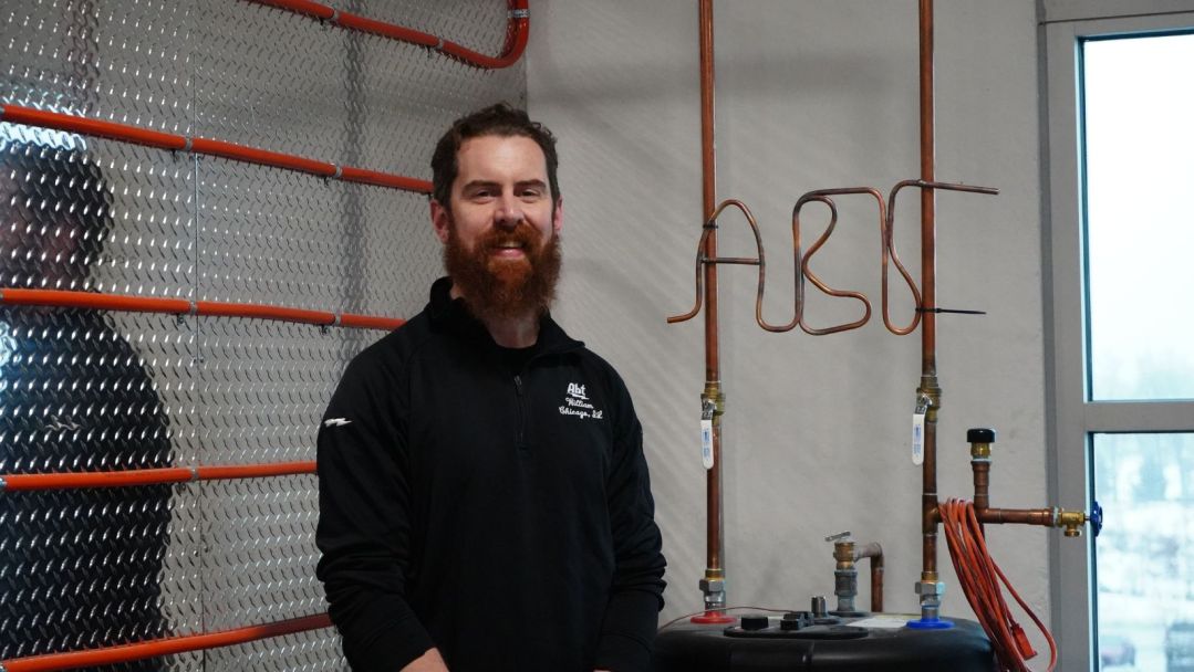 Bill O'Callaghan smiling in our Training Studio beside HVAC training equipment, with a water pipe wire bent to spell "ABT"