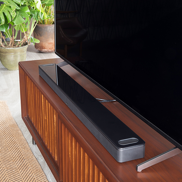 A black Bose Smart Ultra soundbar sitting on a wooden TV stand underneath a flat screen TV