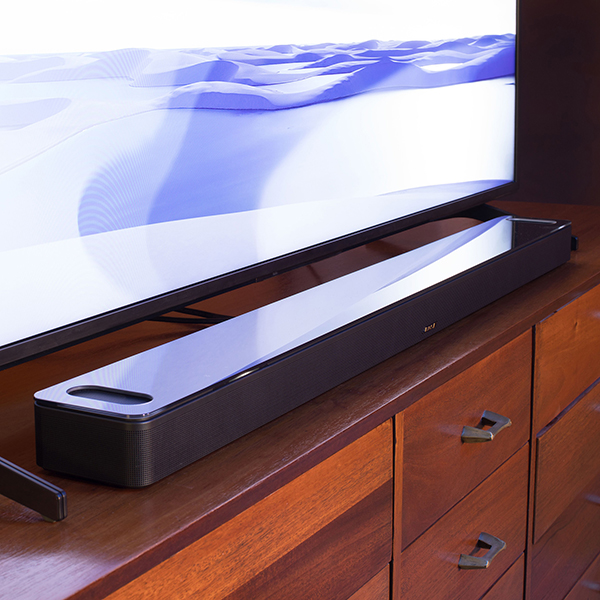 A black Bose Smart Ultra soundbar resting on a wooden TV stand underneath a flat screen TV that is lit up