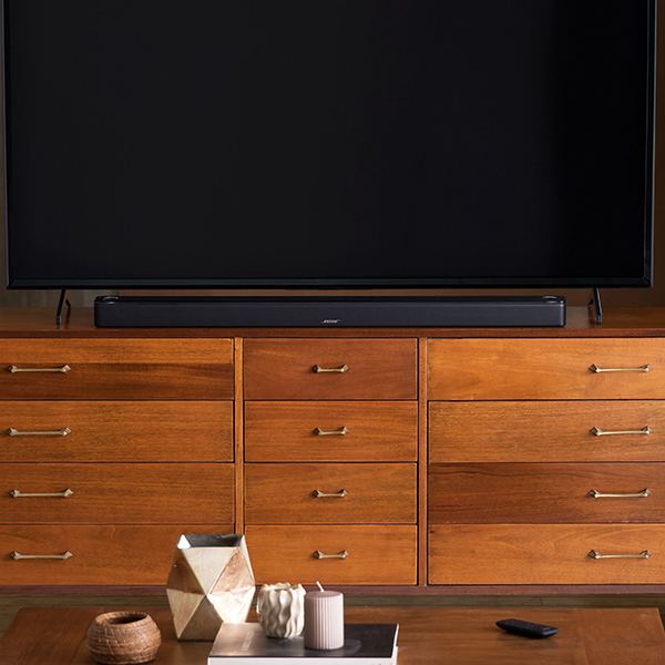 A black Bose Smart Ultra soundbar resting on a wooden TV stand sitting underneath a powered off flat screen TV