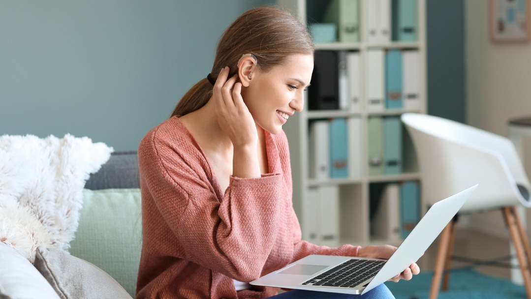 A woman sitting on the couch wearing an over-the-counter hearing aid and looking at a laptop