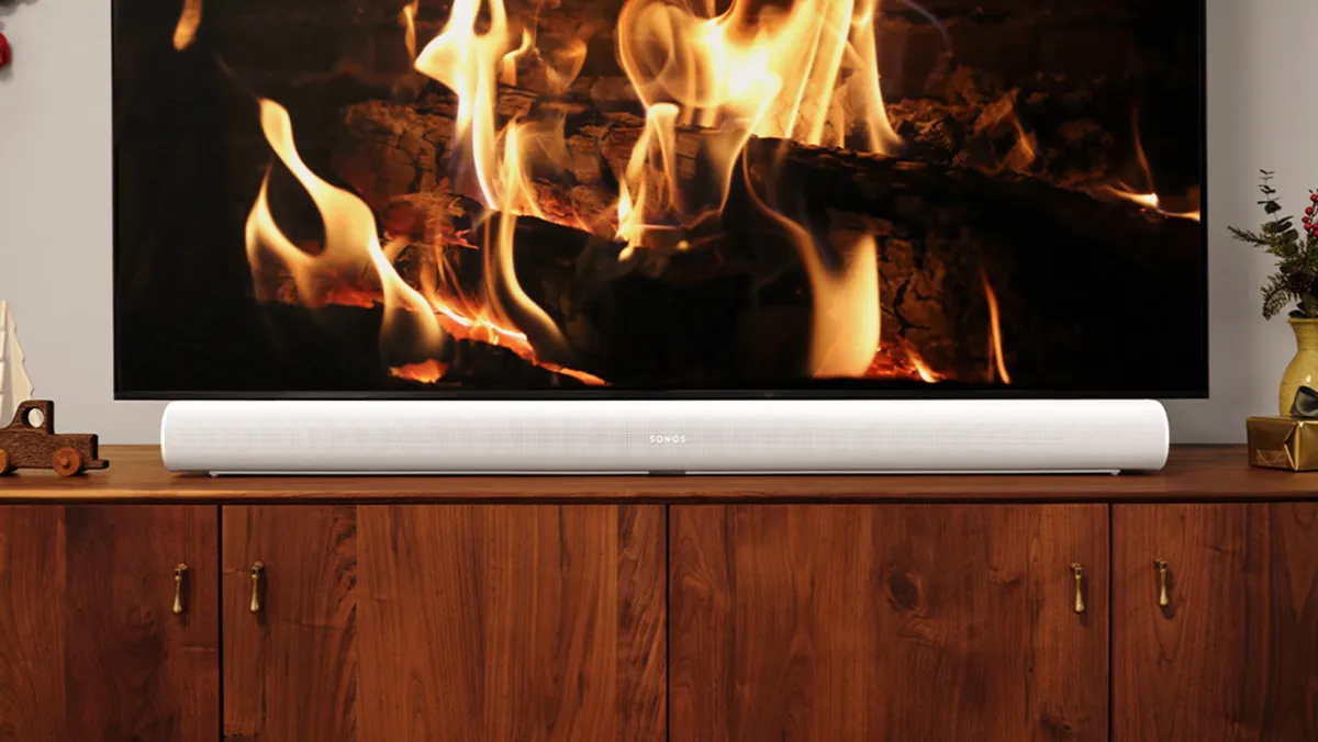 A white Sonos Arc soundbar sitting on a wooden TV stand underneath a flat screen TV that's displaying an image of wood logs burning