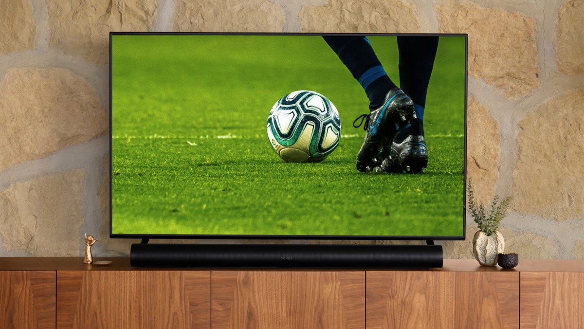A black Sonos Arc soundbar resting on a wooden TV stand underneath a flat screen TV that's displaying a soccer game