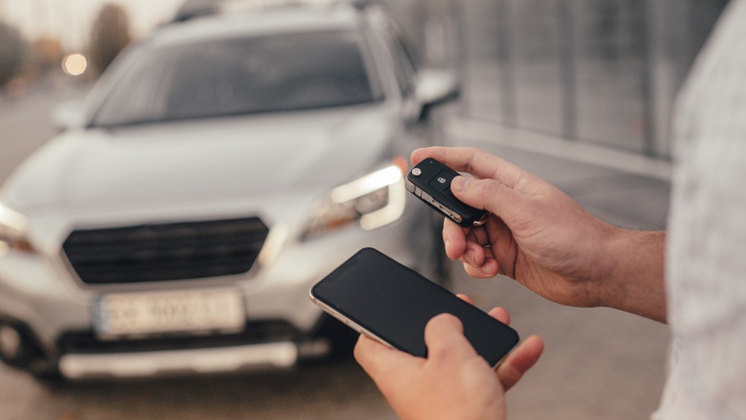 A man holding a remote start key fob along with his smartphone standing in front of a silver car
