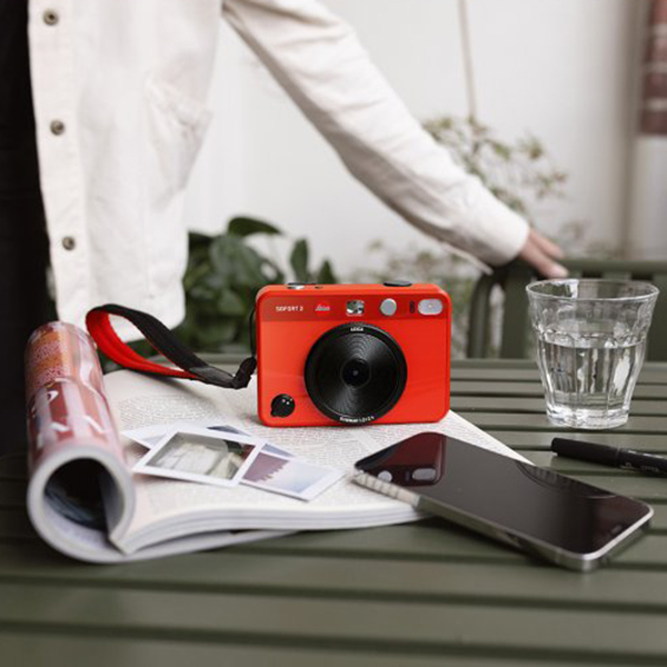 A red instant film camera sitting on a table with a notebook, printed photos and a smartphone.