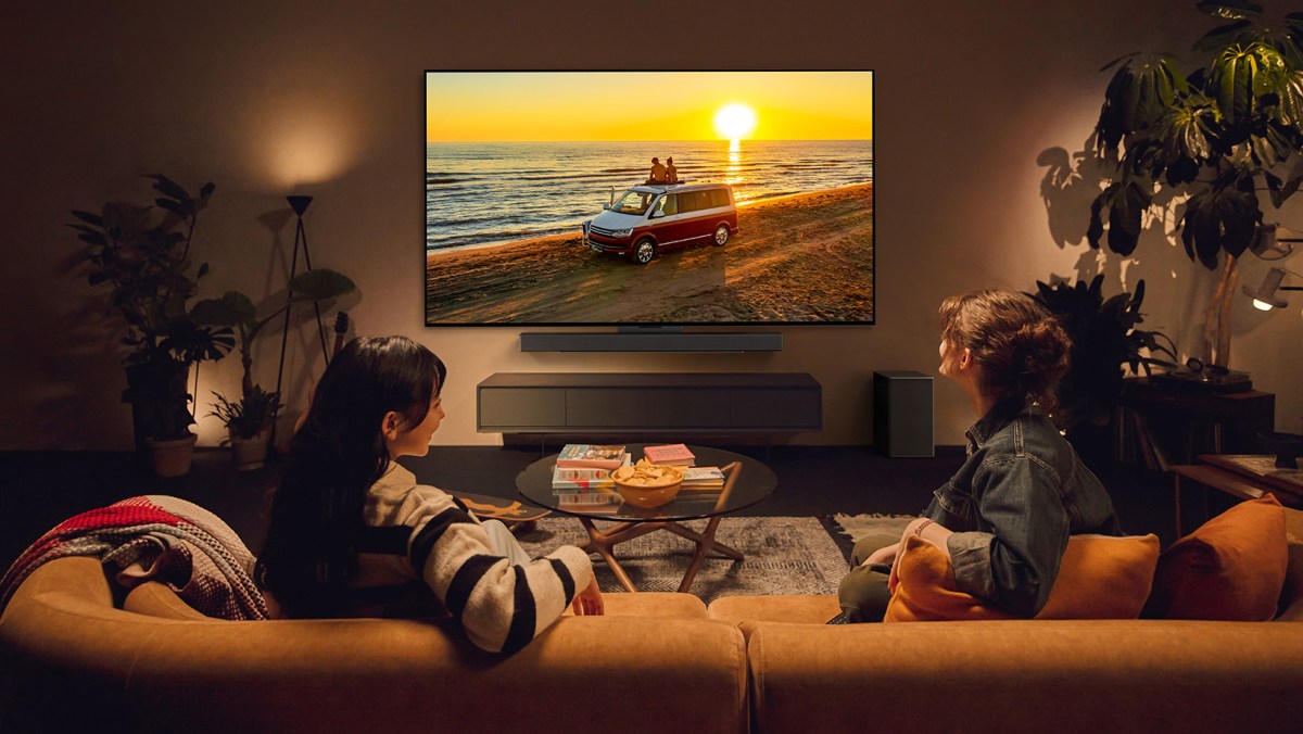Two women sitting on a couch in a living room watching the LG C4, which is displaying a scene of two people sitting on top of a van in front of a shining sunset.