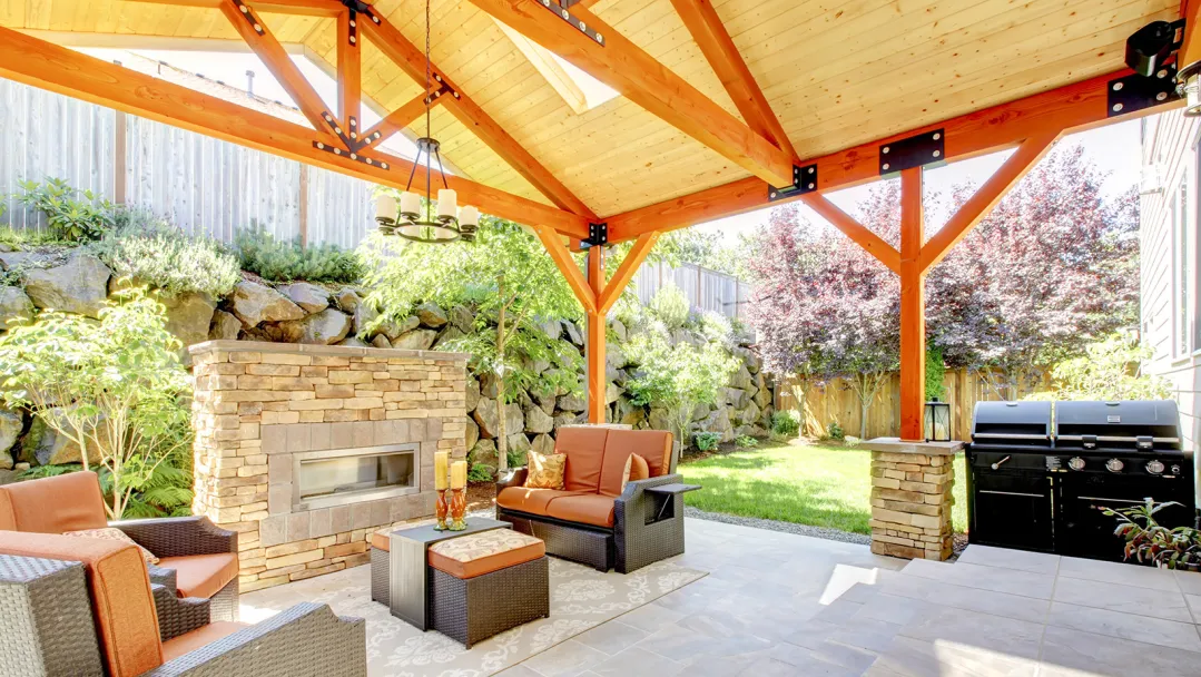 A backyard patio with stone tiles, a wooden awning, outdoor couches, an outdoor table, an outdoor grill and a fireplace.