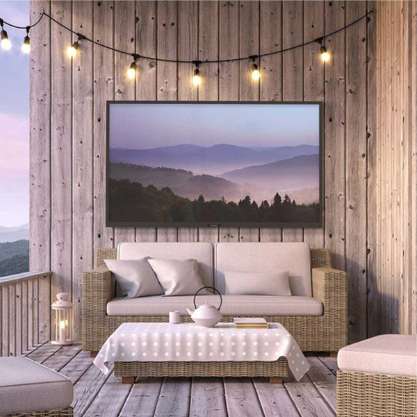 A wooden patio with an outdoor couch, an outdoor table, string lights and an outdoor TV displaying a mountain range.