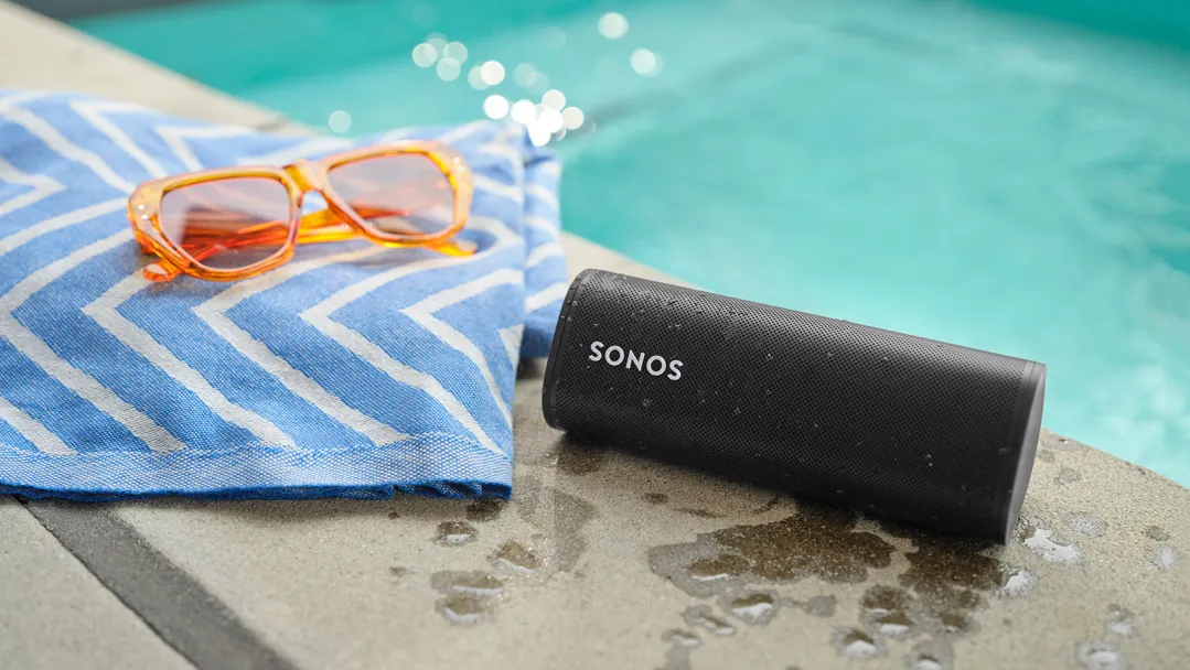 A black Sonos waterproof speaker sitting in front of a pool, with a towel and a pair of swimming goggles next to it.